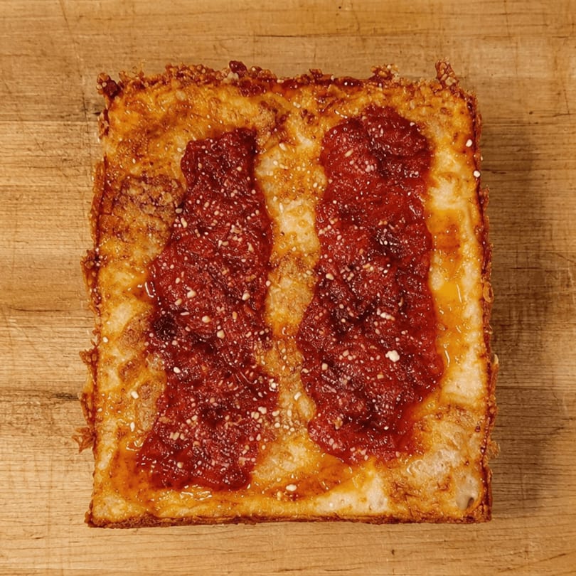 Square Detroit-style pizza with two stripes of red sauce and grated cheese on a wooden surface.