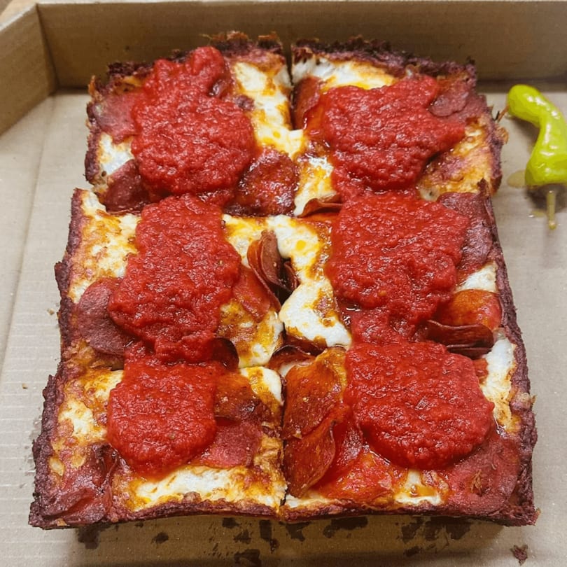 Square Detroit-style pizza with pepperoni and dollops of red sauce in a cardboard box.